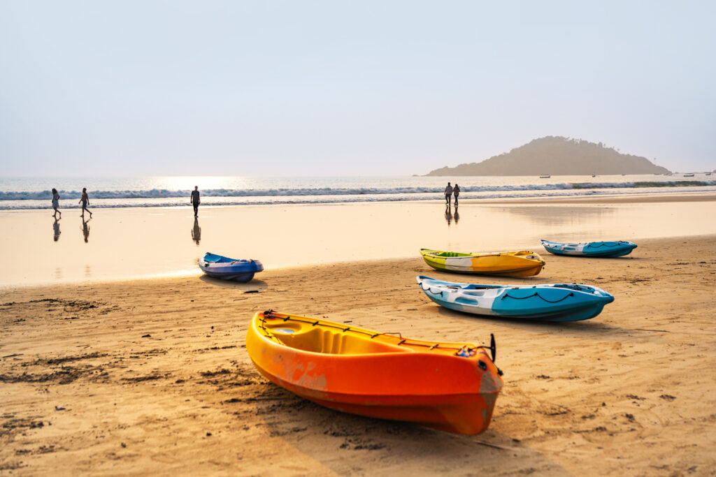 Colorful kayaks on a sandy beach at sunset with people walking along the shoreline in Goa, India.
